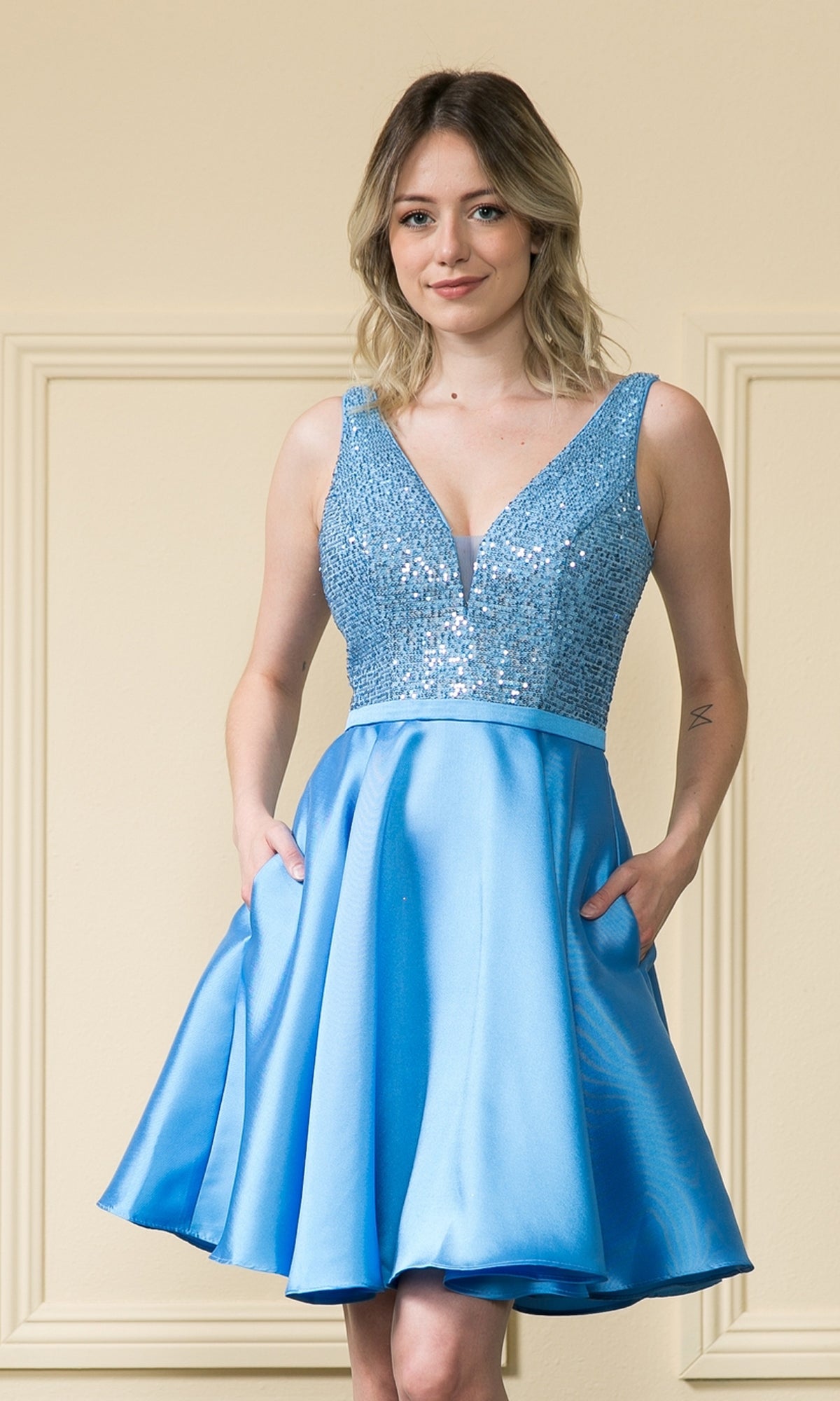 A woman wearing the A-Line Short Homecoming Dress with Pockets - 8954, featuring an embellished bodice, deep V-neckline, and satin knee-length skirt, stands against a beige background.